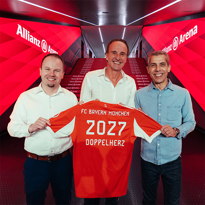 Na fotografiji su tri muškarca koji drže nogometni dres s natpisom "FC Bayern München 2027 DOPPELHERZ" u Allianz Areni. Pozadina je prožeta crvenim svjetlom, stvarajući ambijent vezan uz nogometni klub.