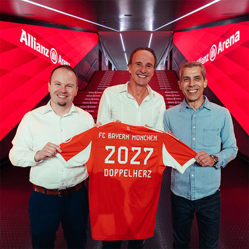 Na fotografiji su tri muškarca koji drže nogometni dres s natpisom "FC Bayern München 2027 DOPPELHERZ" u Allianz Areni. Pozadina je prožeta crvenim svjetlom, stvarajući ambijent vezan uz nogometni klub.
