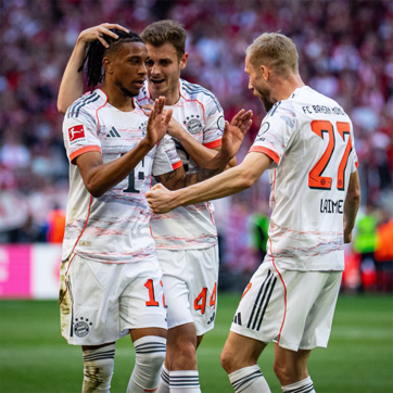 Igrači nogometne momčadi Bayern Munchen slave postignuti gol. Tri igrača u bijelim dresovima međusobno se raduju, a jedan igrač dobiva podršku jednog suigrača koji mu dodiruje glavu. Atmosfera je vesela i uzbuđena.